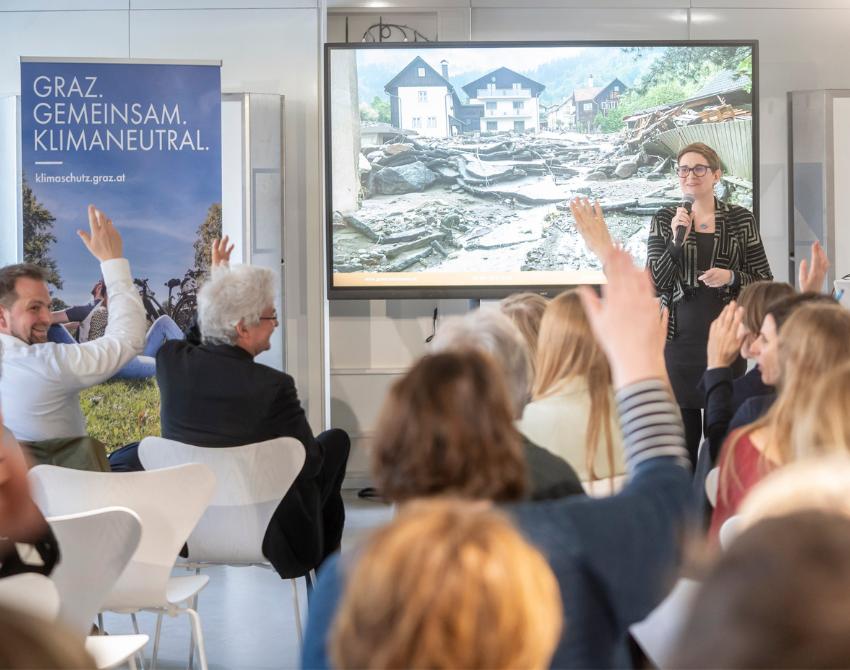 Anna Strobach vor einem Screen mit einem Foto einer Flutkatastrophe. Publikum von hinten sichtbar, einige zeigen auf.