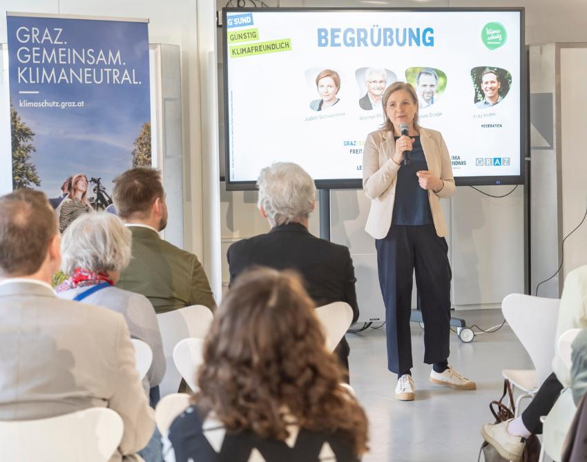 Vizebürgermeisterin Judith Schwentner vor einem Screen auf dem Begüßung steht, spricht in ein Mikrophon. Neben ihr ein Rollup des Referat für Klimaschutzkoordination und Förderungen.