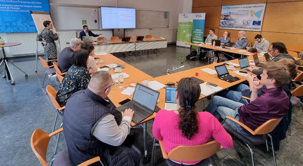 Teilnehmer:innen des Projektteam-Meetings sitzen an Tischen in U-Form, 1 Person steht vor großem Screen und erklärt