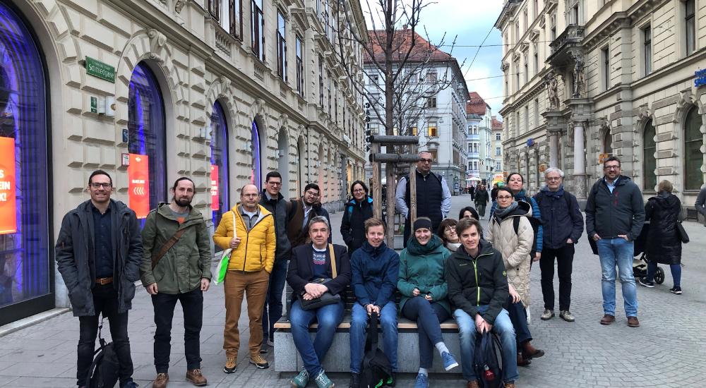 Gruppenfoto des internationalen Ready4Heat-Projektteams in einer Fußgängerzone vor einem kleinen Baum, auf Sitzbänken und im Stehen.