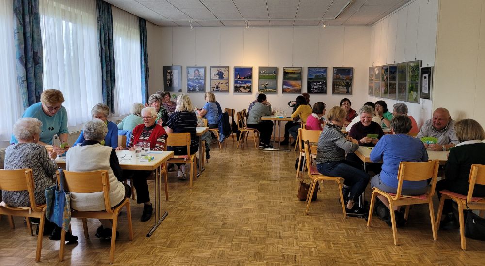 Gesamtansicht eines Raus mit Tischen, and denen Senior:innen sitzen. Im Hintergrund hängen Bilder an der Wand. An der linken Raumseite sind Fenster und Vorhänge.