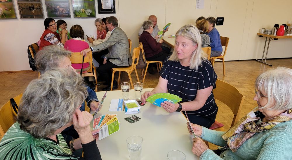 Je vier Senior:innen sitzen an zwei Tischen, Karten in der Handj, hinter ihnen Bilder an der Wand.