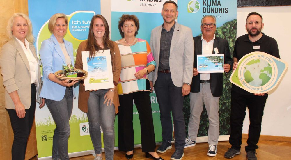 Gruppenbild mit 7 Personen vor Roll-Ups. Eine Person hat eine Klimabündnis-Tafel in der Hand, eine weitere eine Urkunde