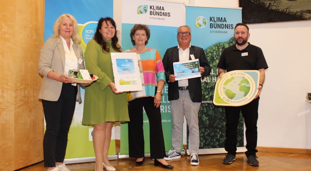 Gruppenbild mit 5 Personen vor Roll-Ups. Eine Person hat eine Klimabündnis-Tafel in der Hand, eine weitere eine Urkunde