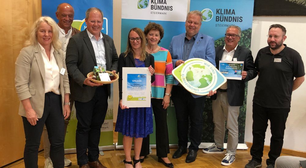 Gruppenbild mit 8 Personen vor Roll-Ups. Eine Person hat eine Klimabündnis-Tafel in der Hand, eine weitere eine Urkunde