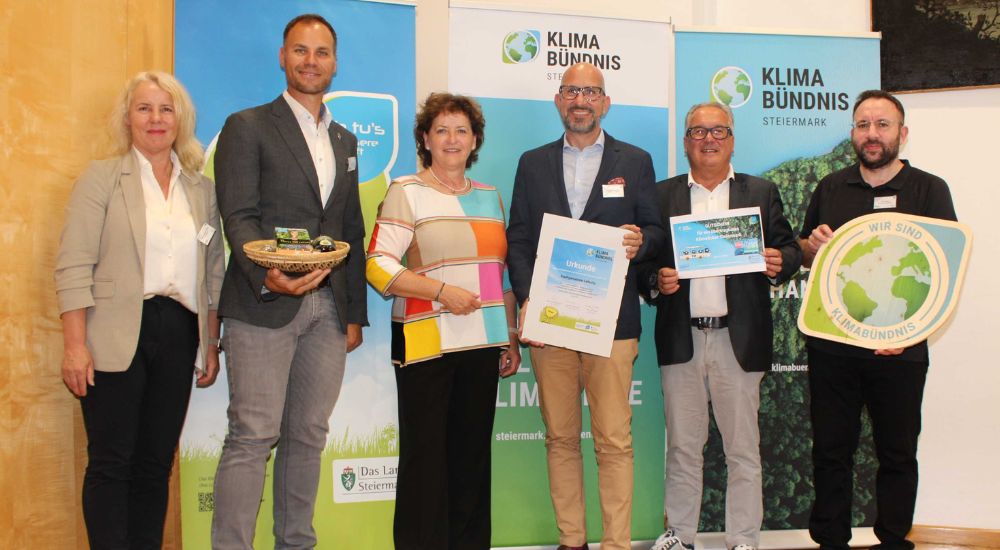 Gruppenbild mit 6 Personen vor Roll-Ups. Eine Person hat eine Klimabündnis-Tafel in der Hand, eine weitere eine Urkunde