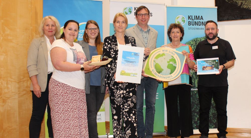 Gruppenbild mit 7 Personen vor Roll-Ups. Eine Person hat eine Klimabündnis-Tafel in der Hand, eine weitere eine Urkunde