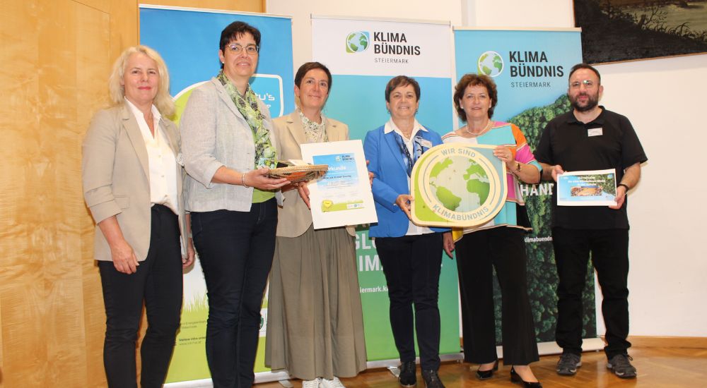 Gruppenbild mit 6 Personen vor Roll-Ups. Eine Person hat eine Klimabündnis-Tafel in der Hand, eine weitere eine Urkunde