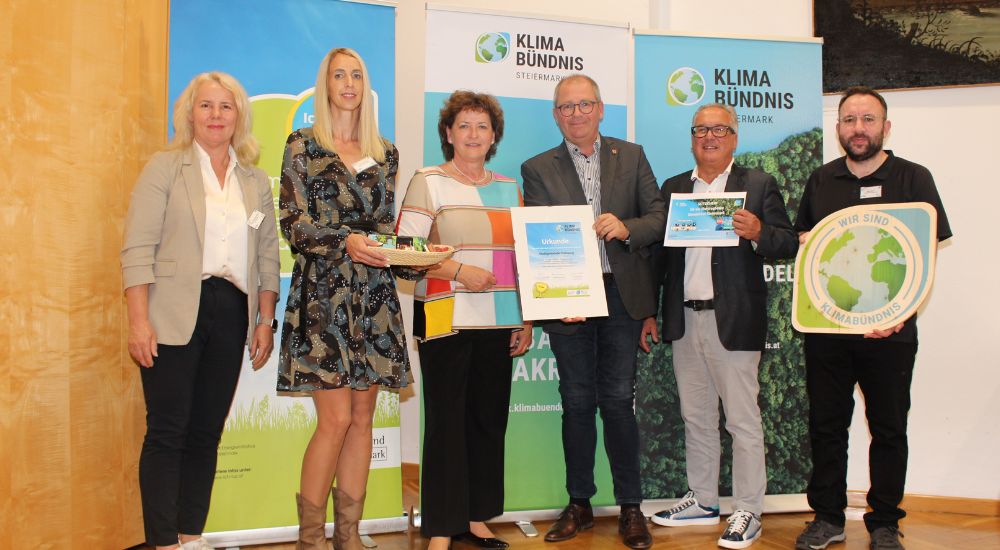 Gruppenbild mit 6 Personen vor Roll-Ups. Eine Person hat eine Klimabündnis-Tafel in der Hand, eine weitere eine Urkunde