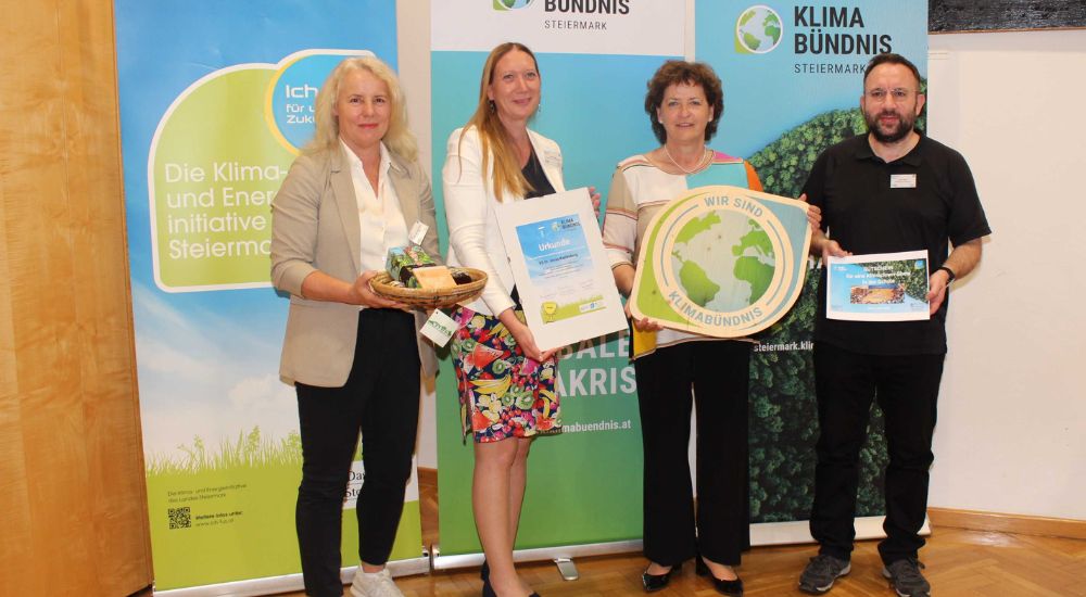 Gruppenbild mit 4 Personen vor Roll-Ups. Eine Person hat eine Klimabündnis-Tafel in der Hand, eine weitere eine Urkunde