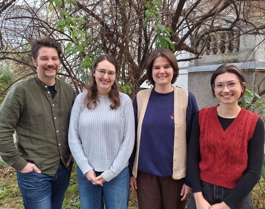 Vier Personen mittleren Alters, links ein mann, dann drei junge Frauen, die in die Kamera lächeln