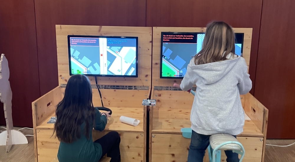 zwei kinder sitzen an interaktiver station, ein kind fährt mit einem auto, das andere auf einem fahrrad