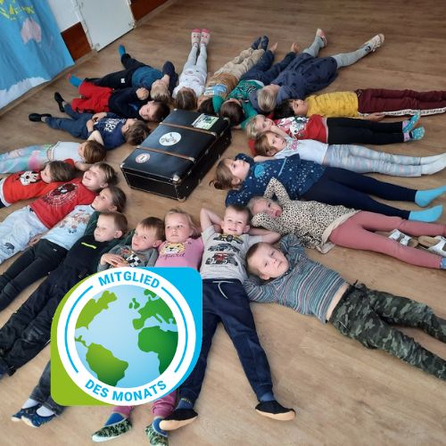 Kinder liegen am Boden rund um einen Koffer mit Klimabündnis-Logo und Mitglied des Monats Beschriftung links unten