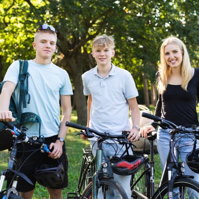 drei Jugendliche, zwei Jungs und ein Mädchen stehen mit ihren Rädern im Grünen und lachen in die Kamera